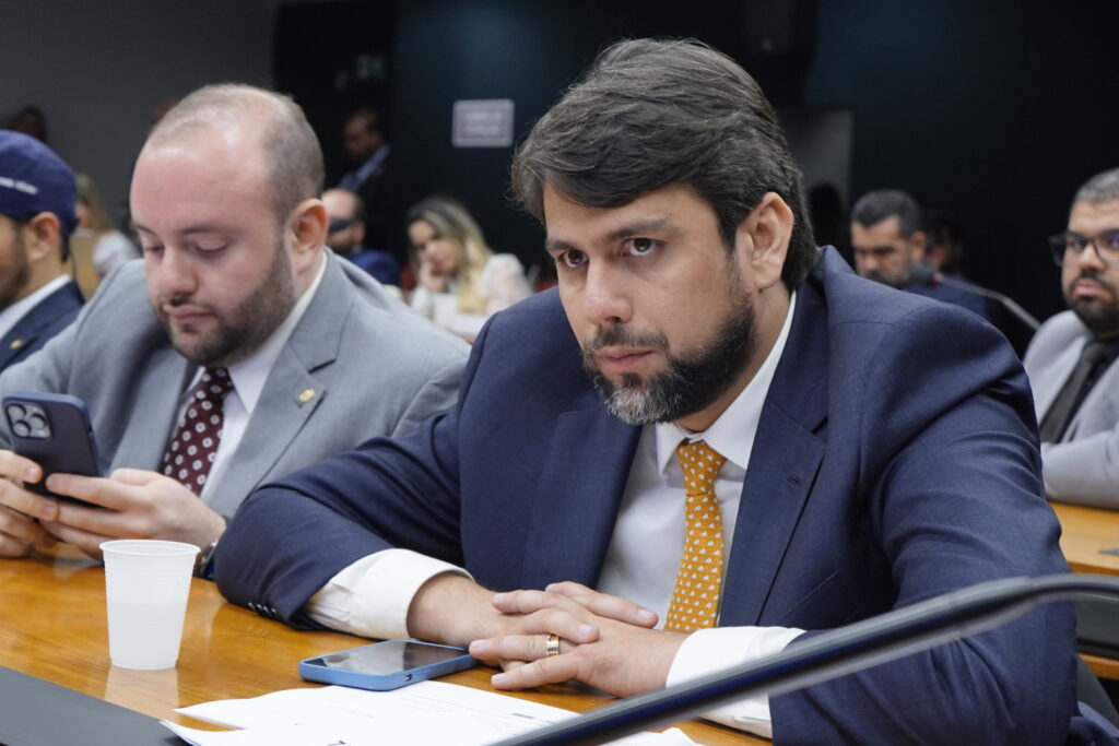 Foto com o deputado Pedro Lucas Fernandes em destaque com as mãos cruzadas apoiadas sobre a mesa. Ao fundo aparecem outros parlamentares.